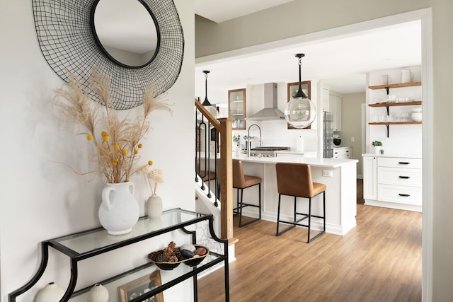 kitchen with open shelves, wall chimney range hood, a kitchen bar, light wood-type flooring, and white cabinetry