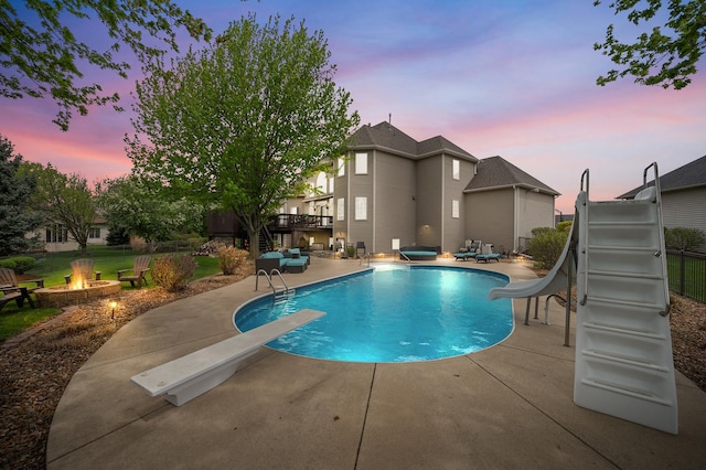 pool at dusk with an outdoor fire pit, a diving board, a water slide, a patio area, and an outdoor pool