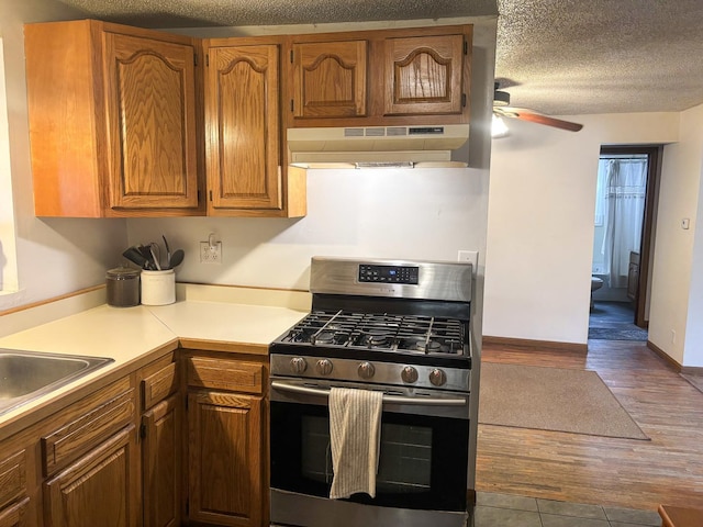 kitchen featuring gas range, ceiling fan, under cabinet range hood, brown cabinets, and dark wood-type flooring