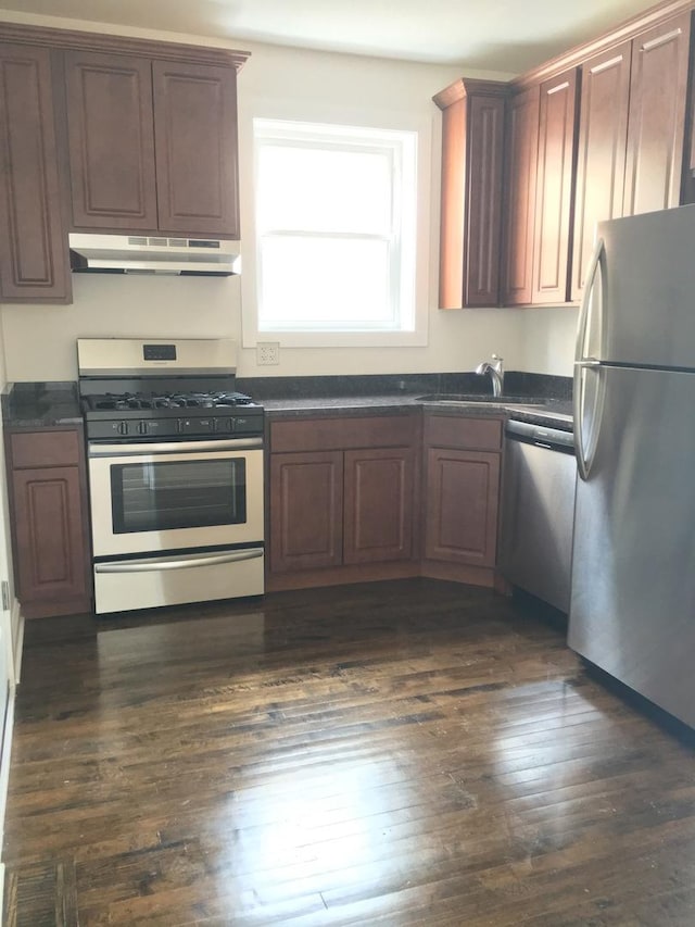 kitchen featuring dark wood-style flooring, dark countertops, a sink, stainless steel appliances, and under cabinet range hood