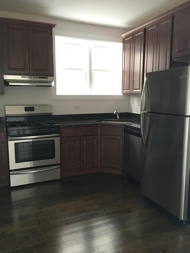 kitchen with dark countertops, appliances with stainless steel finishes, a sink, under cabinet range hood, and dark wood-style floors