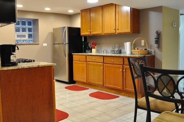 kitchen with brown cabinetry, recessed lighting, light stone countertops, and freestanding refrigerator