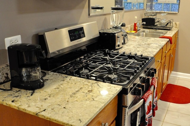 kitchen featuring baseboards, light tile patterned floors, stainless steel gas range oven, brown cabinets, and a sink