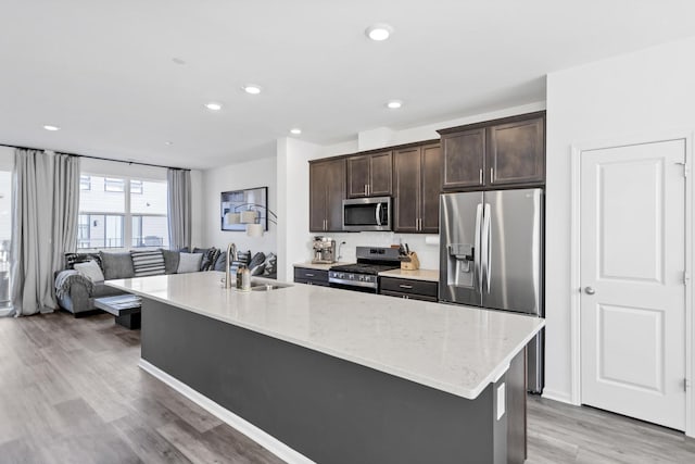 kitchen featuring an island with sink, light stone countertops, dark brown cabinetry, light wood-type flooring, and appliances with stainless steel finishes