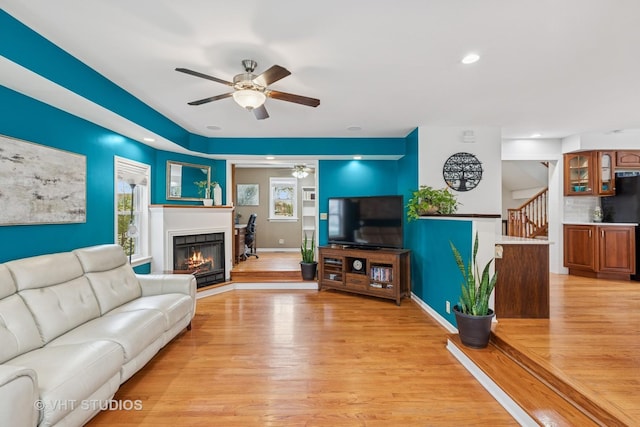 living room with light wood finished floors, a glass covered fireplace, stairs, a ceiling fan, and recessed lighting