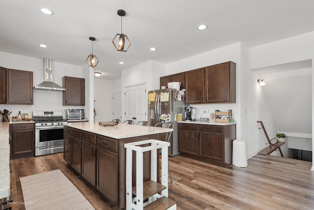 kitchen with stainless steel appliances, wall chimney exhaust hood, dark brown cabinetry, dark wood-style floors, and decorative light fixtures