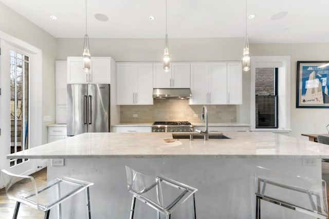 kitchen featuring white cabinets, a sink, under cabinet range hood, high end refrigerator, and backsplash