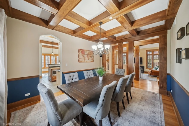 dining area featuring arched walkways, beam ceiling, an inviting chandelier, light wood-style floors, and coffered ceiling