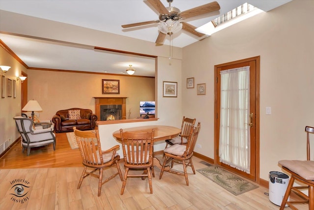 dining space with ceiling fan, a warm lit fireplace, light wood-style floors, a skylight, and baseboards
