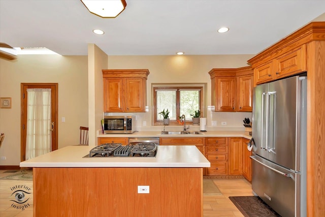 kitchen featuring appliances with stainless steel finishes, a sink, light countertops, a center island, and brown cabinetry