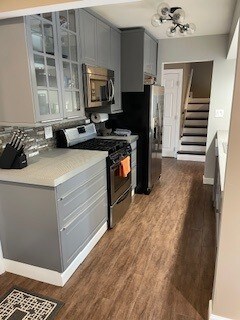 kitchen featuring light countertops, gray cabinets, glass insert cabinets, appliances with stainless steel finishes, and dark wood-style flooring