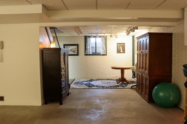 below grade area featuring rail lighting, brick wall, and a drop ceiling