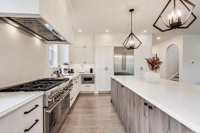 kitchen with recessed lighting, high end appliances, light wood finished floors, wall chimney exhaust hood, and a notable chandelier