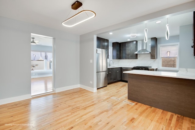 kitchen with freestanding refrigerator, light wood-style flooring, wall chimney range hood, light countertops, and decorative backsplash