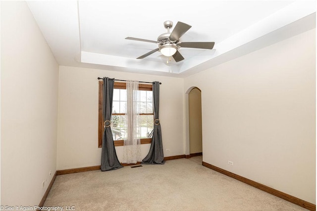 empty room with a ceiling fan, light colored carpet, arched walkways, baseboards, and a tray ceiling