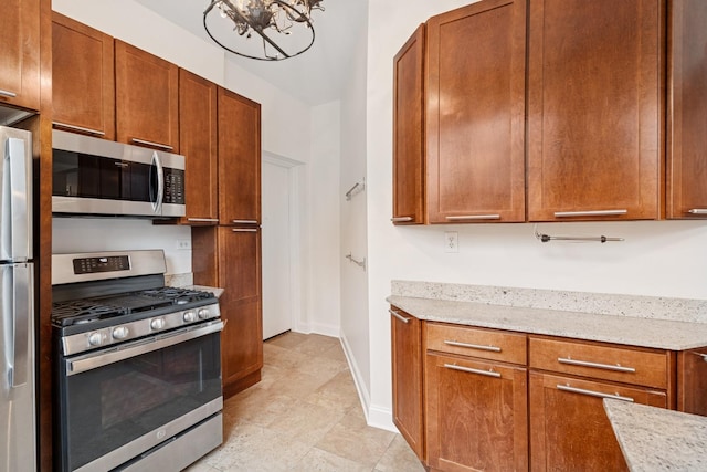 kitchen with light tile patterned floors, an inviting chandelier, appliances with stainless steel finishes, brown cabinets, and light stone countertops