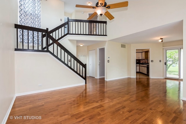 unfurnished living room with stairs, a towering ceiling, wood finished floors, baseboards, and a ceiling fan