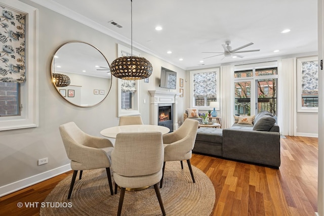 dining space with a ceiling fan, a warm lit fireplace, ornamental molding, wood finished floors, and baseboards