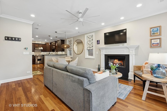 living room with ceiling fan, recessed lighting, a fireplace with flush hearth, light wood-style flooring, and crown molding