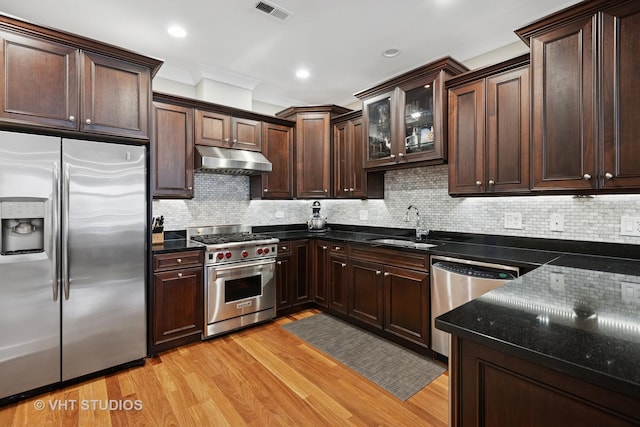 kitchen with stainless steel appliances, under cabinet range hood, light wood-style flooring, dark stone countertops, and a sink