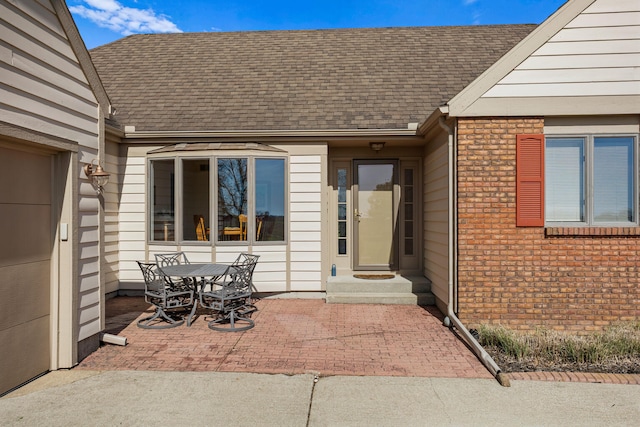 view of exterior entry featuring brick siding, a shingled roof, a patio, and outdoor dining space