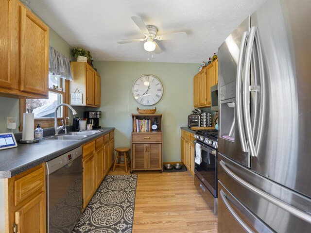 kitchen with a sink, appliances with stainless steel finishes, ceiling fan, dark countertops, and light wood finished floors