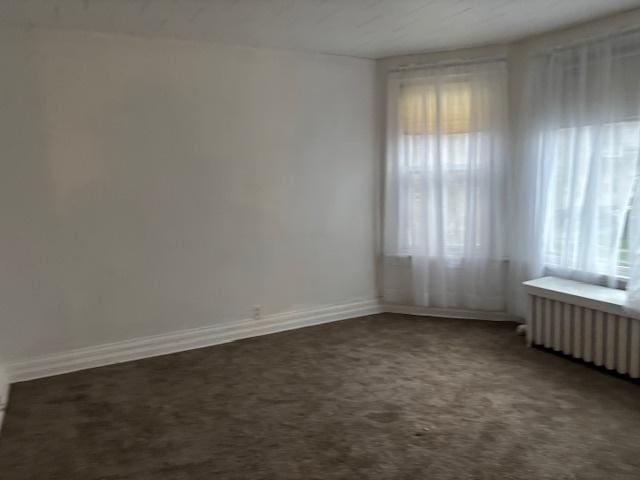 spare room featuring dark carpet, baseboards, and radiator heating unit