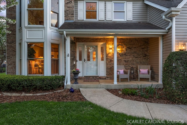 doorway to property featuring a porch, roof with shingles, and brick siding