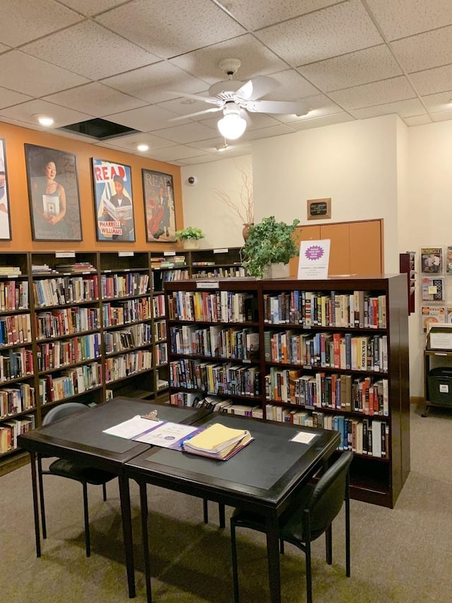 sitting room with wall of books, ceiling fan, a drop ceiling, and carpet floors