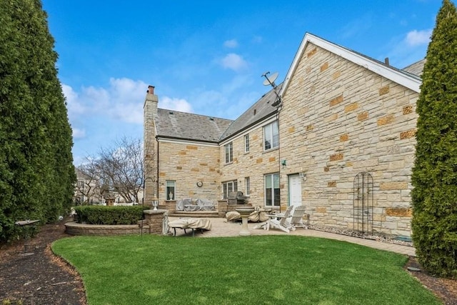 back of property with a high end roof, a yard, a patio area, stone siding, and a chimney