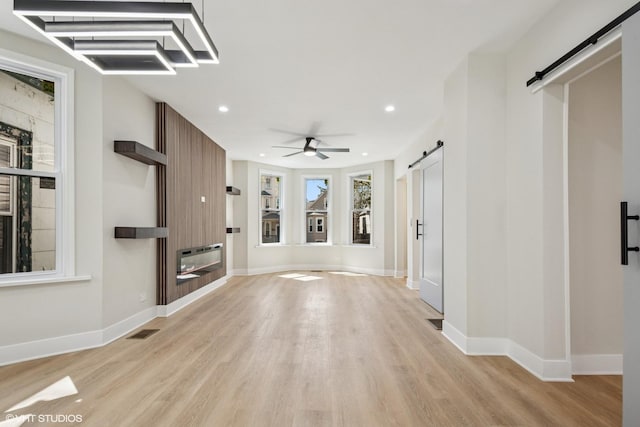 unfurnished living room featuring ceiling fan, a barn door, light wood-style floors, a glass covered fireplace, and visible vents