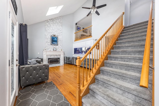 staircase featuring high vaulted ceiling, wood finished floors, a skylight, a fireplace with flush hearth, and ceiling fan
