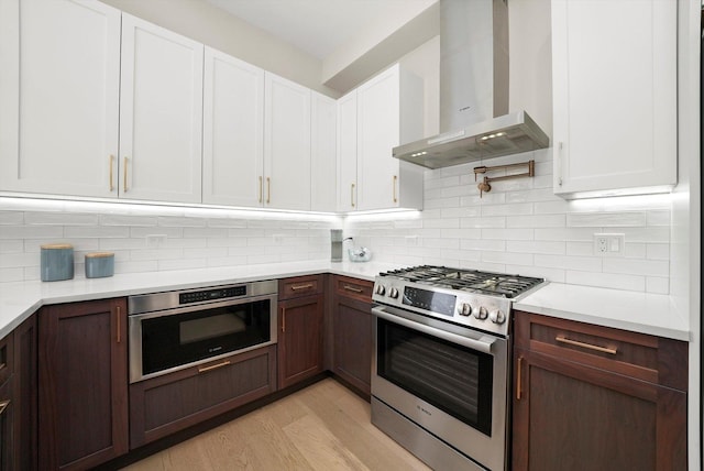 kitchen with light wood-type flooring, wall chimney range hood, white cabinetry, appliances with stainless steel finishes, and light countertops