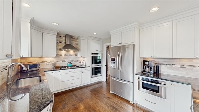 kitchen featuring dark stone counters, decorative backsplash, stainless steel appliances, wall chimney range hood, and a sink