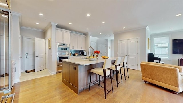 kitchen with a breakfast bar, stainless steel double oven, light wood-type flooring, a center island with sink, and ornamental molding