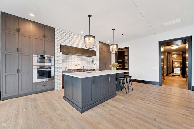 kitchen with baseboards, double oven, an island with sink, light countertops, and light wood-style flooring