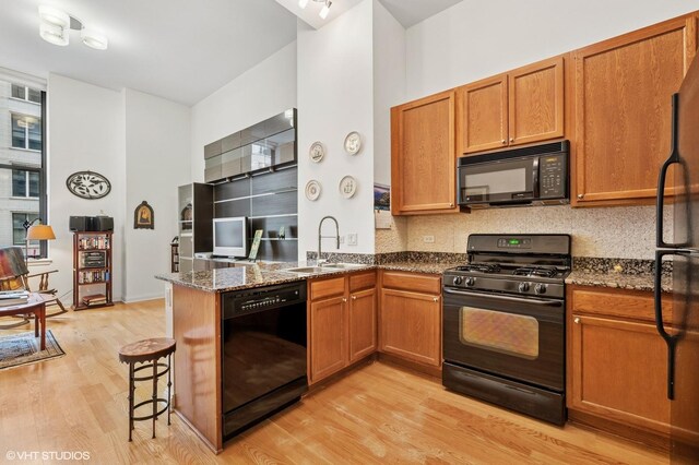 kitchen with light wood-style flooring, brown cabinetry, a sink, dark stone countertops, and black appliances