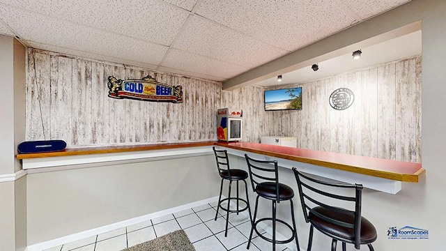 bar with baseboards, wooden walls, a paneled ceiling, tile patterned floors, and a dry bar