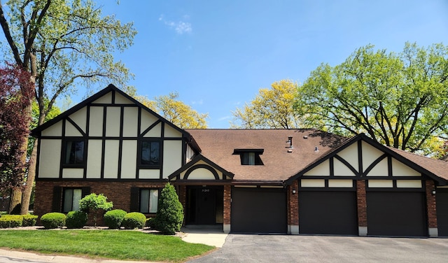 tudor house with brick siding, an attached garage, driveway, a front lawn, and roof with shingles