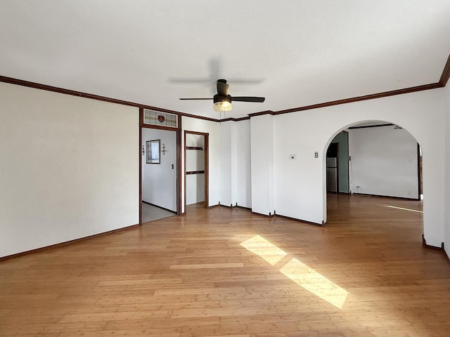 empty room featuring a ceiling fan, crown molding, light wood-style floors, arched walkways, and baseboards