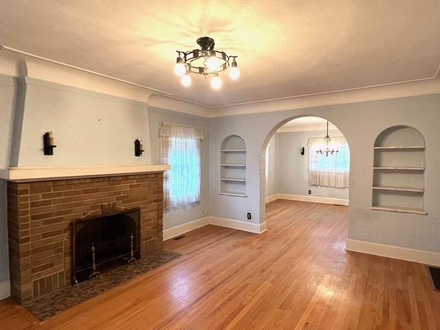 unfurnished living room with wood finished floors, built in features, baseboards, a notable chandelier, and a brick fireplace