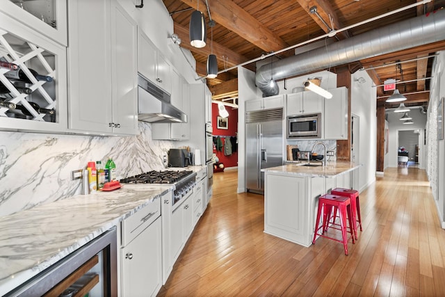 kitchen with beverage cooler, under cabinet range hood, a sink, built in appliances, and a breakfast bar area