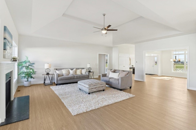 living area featuring light wood-style flooring, a tray ceiling, baseboards, a fireplace with raised hearth, and a ceiling fan