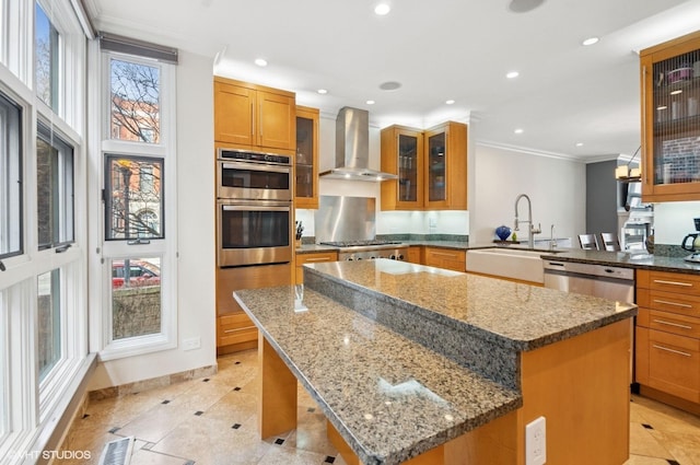 kitchen featuring wall chimney exhaust hood, a sink, appliances with stainless steel finishes, a kitchen island, and ornamental molding