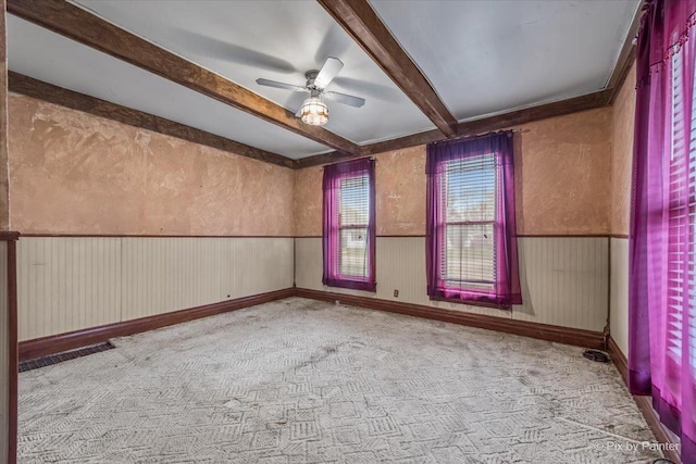 carpeted empty room featuring beam ceiling, a wainscoted wall, and a ceiling fan