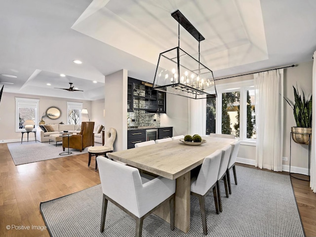 dining area featuring dark wood finished floors, a raised ceiling, ceiling fan with notable chandelier, baseboards, and bar