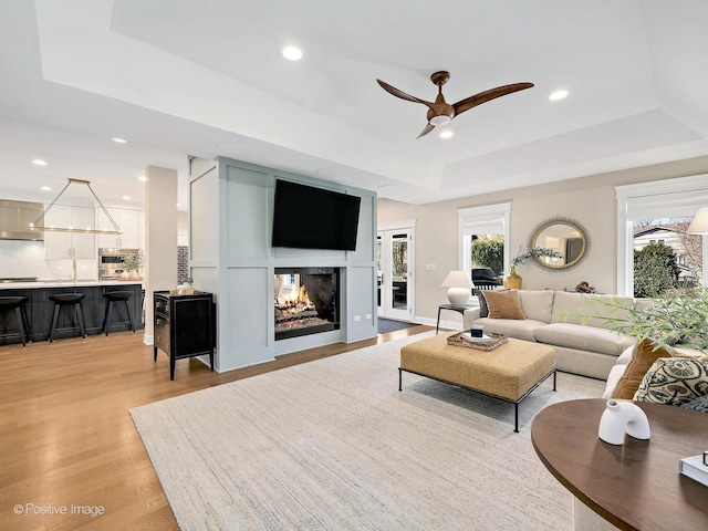 living area featuring a multi sided fireplace, light wood-type flooring, ceiling fan, a raised ceiling, and recessed lighting