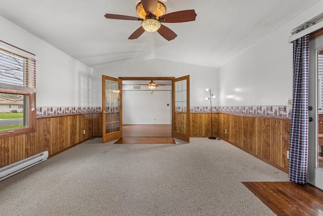 carpeted empty room with wood walls, vaulted ceiling, a ceiling fan, a baseboard heating unit, and wainscoting