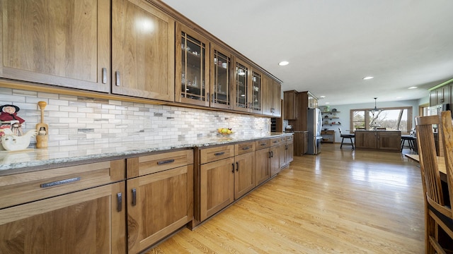 kitchen featuring light wood finished floors, tasteful backsplash, freestanding refrigerator, glass insert cabinets, and light stone countertops