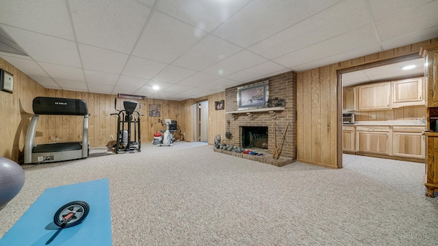workout area with wooden walls, a brick fireplace, carpet flooring, and a drop ceiling